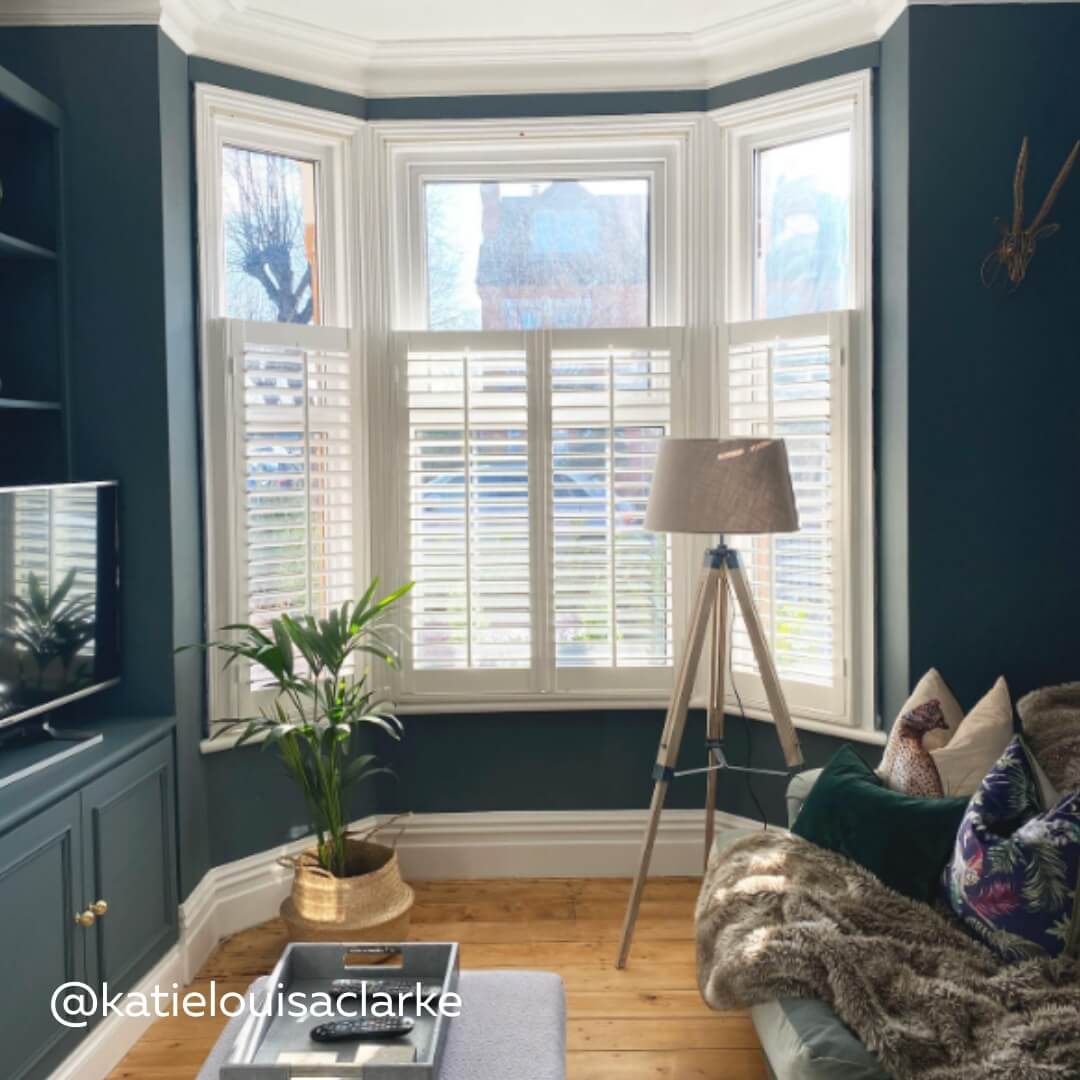 White Cafe Style Shutters with central tilt rod in living room against blue walls and a tripod lamp